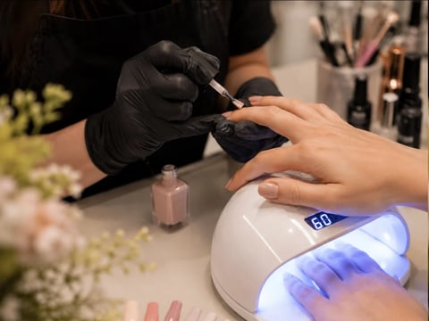Manicurist in black gloves applying nail polish to clients hand under UV lamp in salon setting